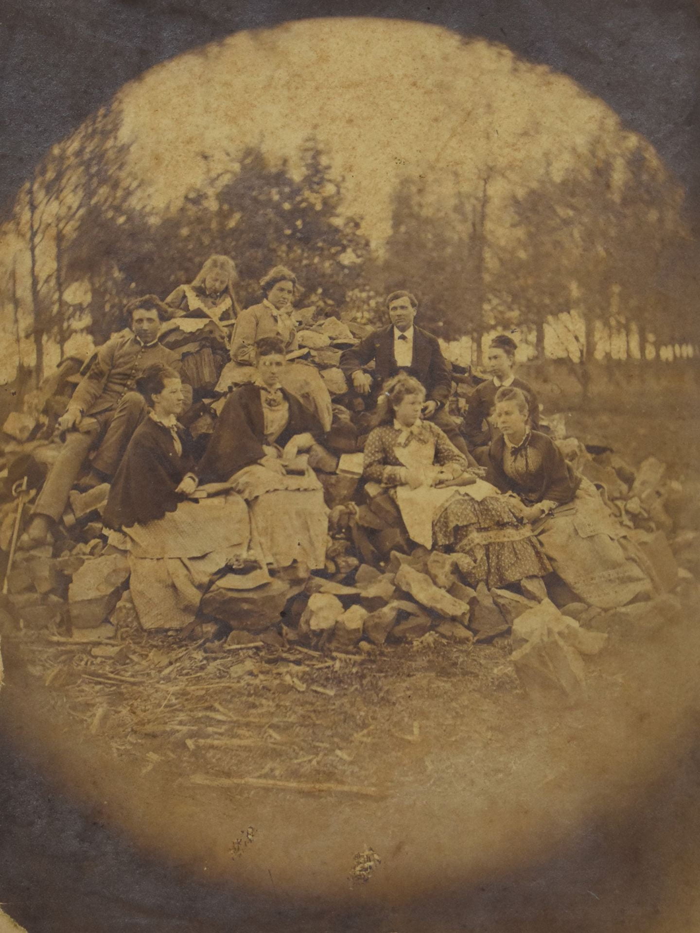 A black and white posed portrait with nine people sitting on a pile of rocks outside with trees in the background. They are all dressed formally in 19th century dresses and suits.