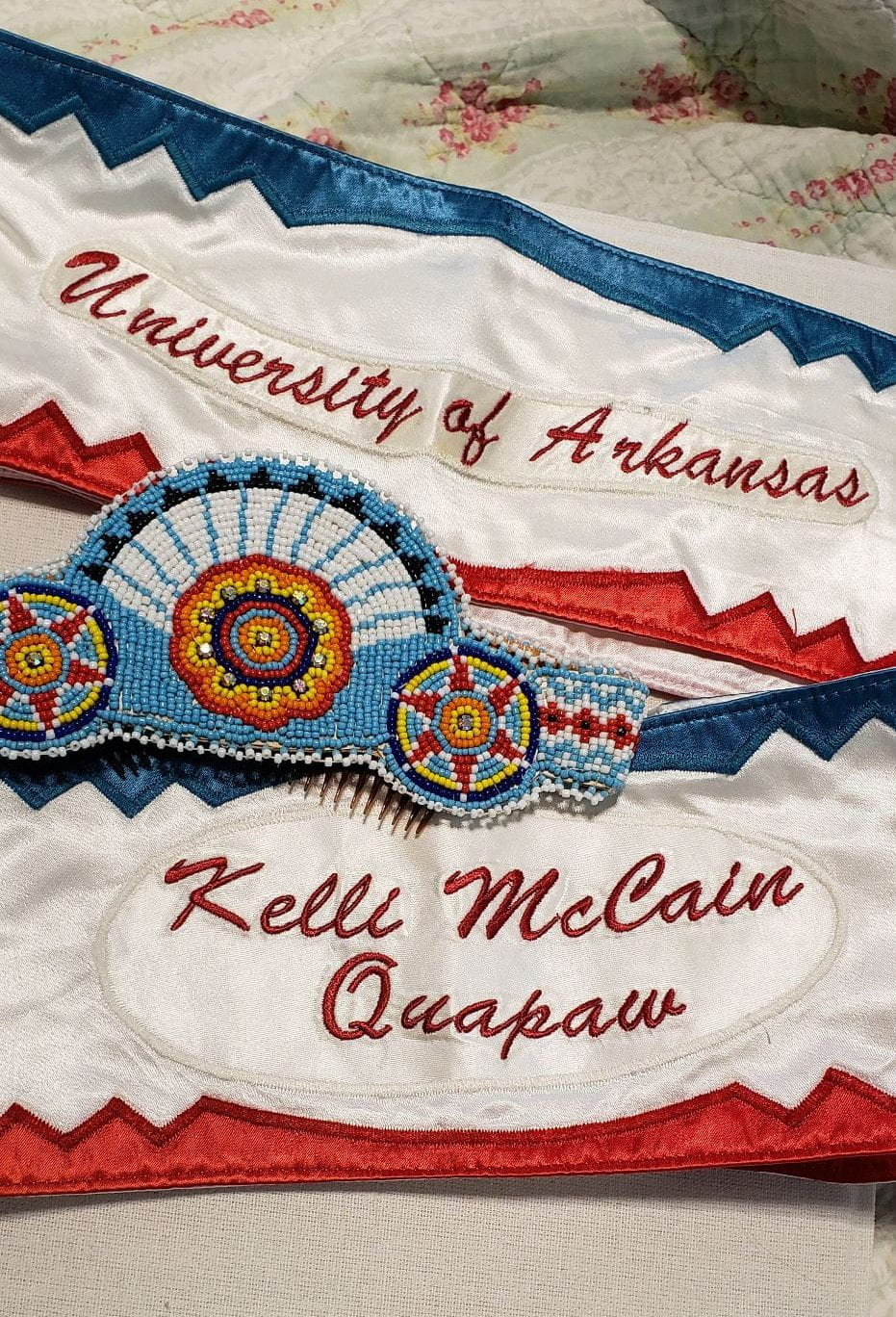 PowPow regalia laid out, including two sashs bordered with blue at the top, red at the bottom, and white in the middle. One says "University of Arkansas" and the other says "Kelli McCain Quapaw." There is also an intricately beaded crown with a blue background and three circular designs.