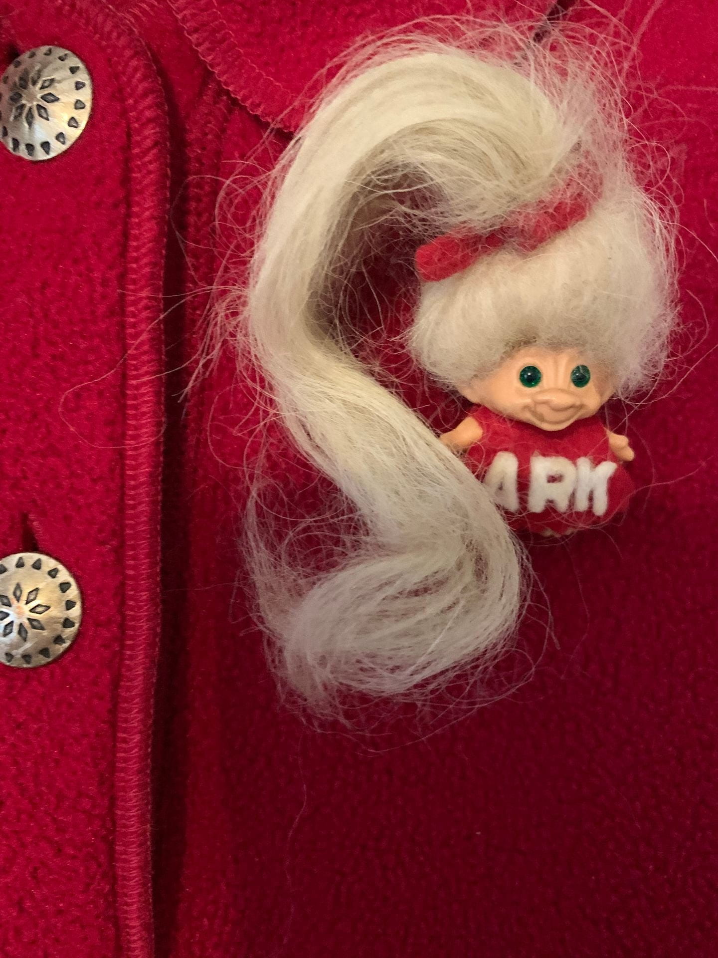 A small troll doll with long blonde hair held in a ponytail with a red bow. The doll wears a red dress that says 