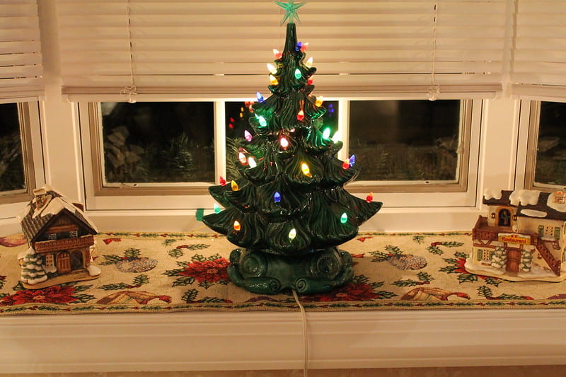 A one foot tall decorative ceramic in the shape of a pine tree. It has colorful lines attached to it and sits on a windowsill with two small ceramic house figurines decorated with snow.
