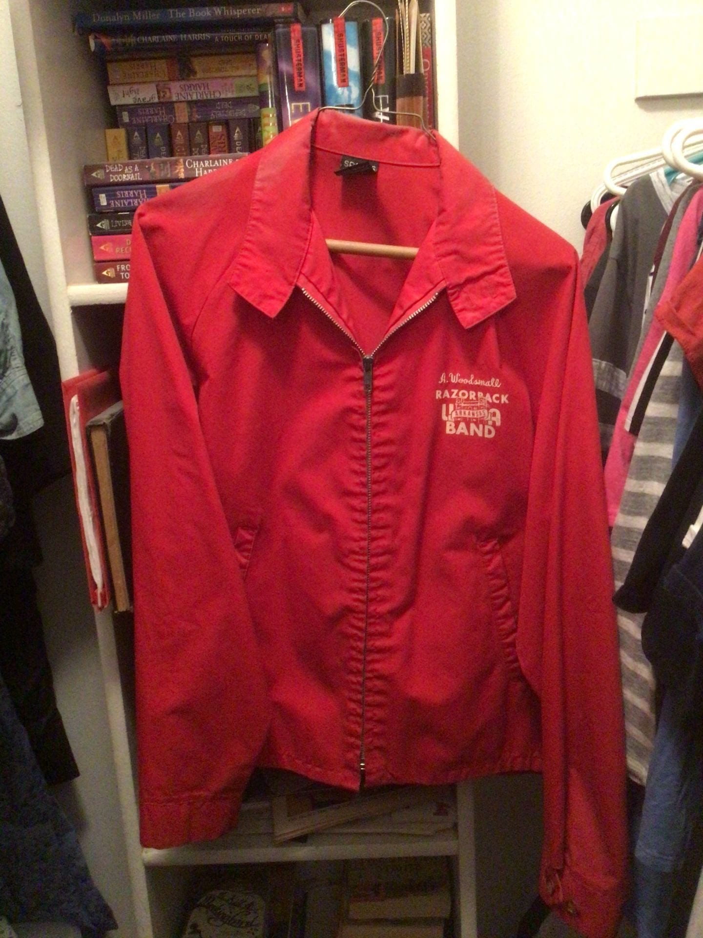 Red jacket with letters engraved in white on left side that states "A. Woodsmall UA Razorback Band." It is on a hanger in a closet and includes side pockets and a zipper.