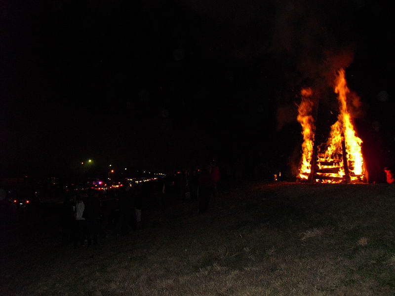 A lowlit outdoor night scene with a very large bonfire lit on a raised grassy area to the right, while a crowd of people stand next to it and cars drive by.