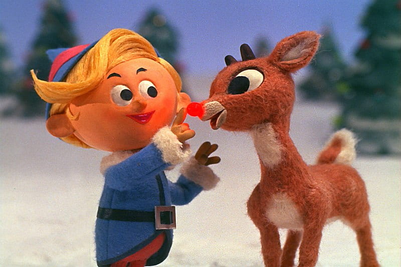 A cartoon elf touching the red nose of a reindeer next to them. A winter scene of snow and evergreen trees is behind them. The figures are three dimensional and look to be made of clay.