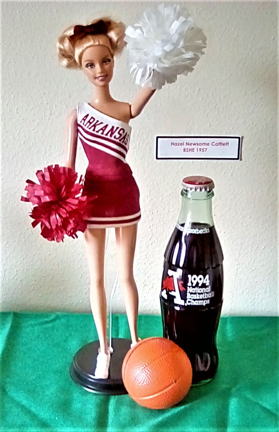 A cheerleader Barbie with one red and one white pom-pom in her hands. She wears a red and white outfit that says "Arkansas" on it. There is a small 1994 National Basketball Champs Coca-Cola bottle and small plastic basketball next to the doll.