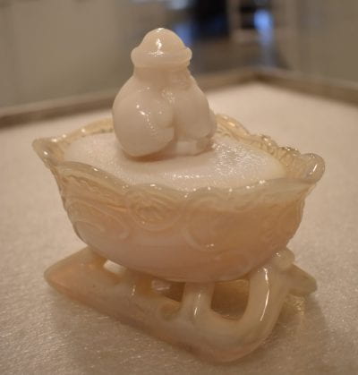 A small white ceramic in the shape of a sleigh with a man in it. The man is wearing a coat, hat, and has a long beard.