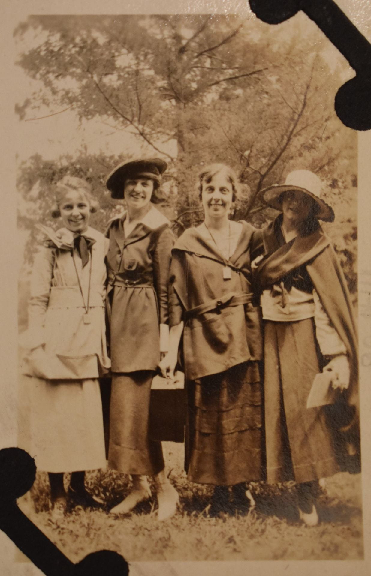 A black and white image of four women posing next to each other. They are wearing early 20th century dresses and hats and smiling at the camera.