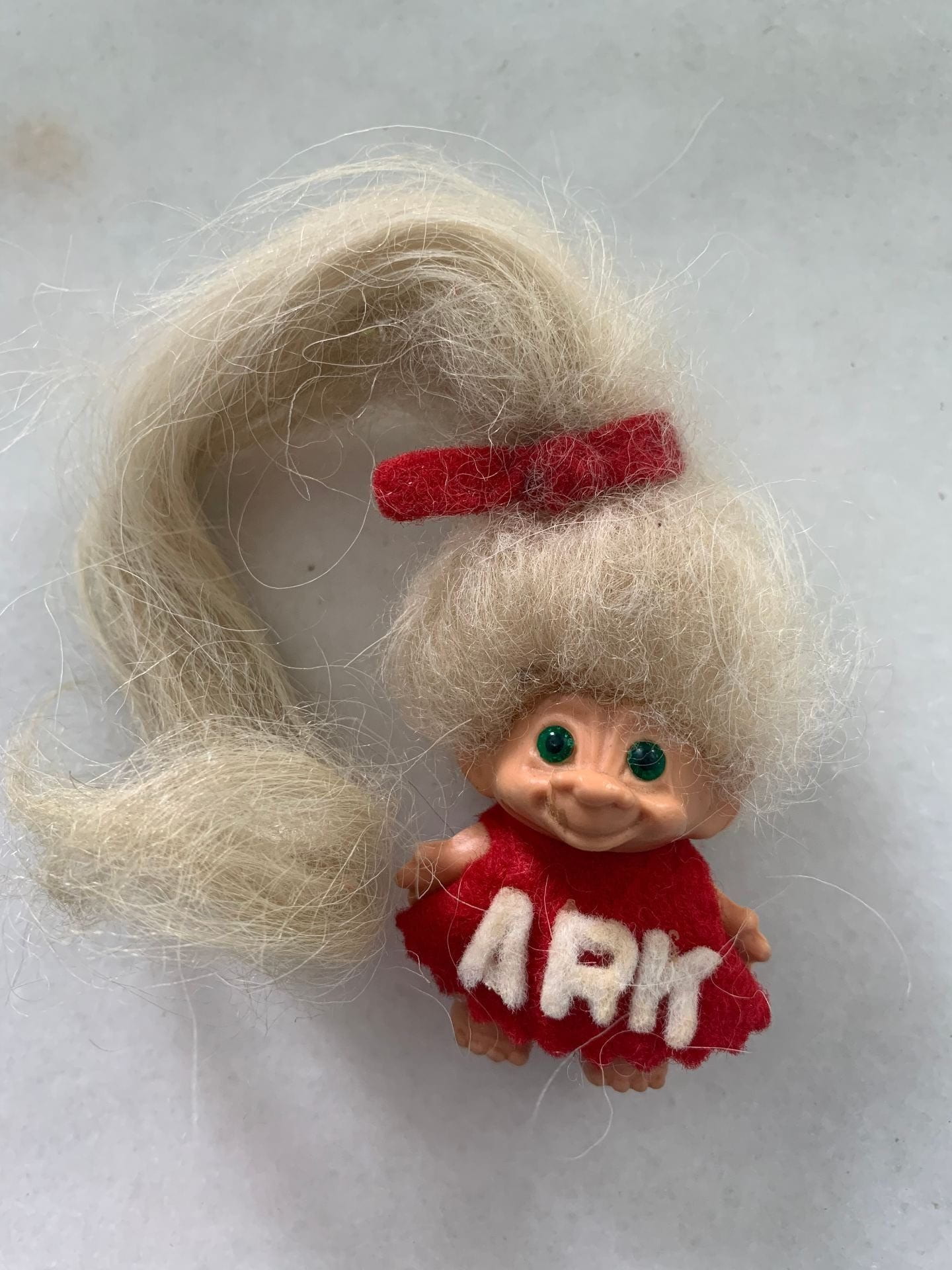 A small troll doll with long blonde hair held in a ponytail with a red bow. The doll wears a red dress that says "ARK" across it.