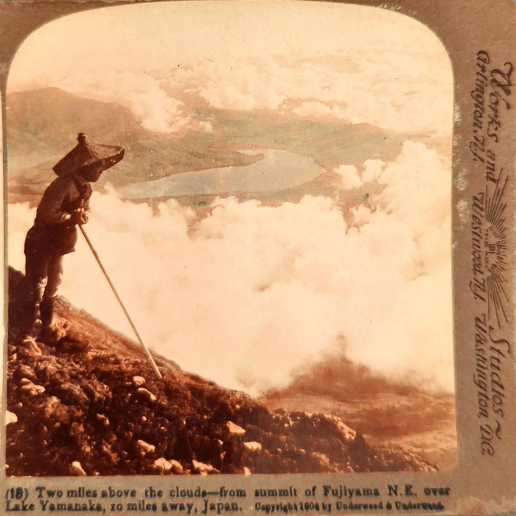 A black and white stereograph of a man wearing a large hat looking at the landscape above the clouds. At the bottom, the image is titled "Two miles above the clouds - from summit of Fujiyama N.E. over Lake Yamanaka, 10 miles away, Japan."