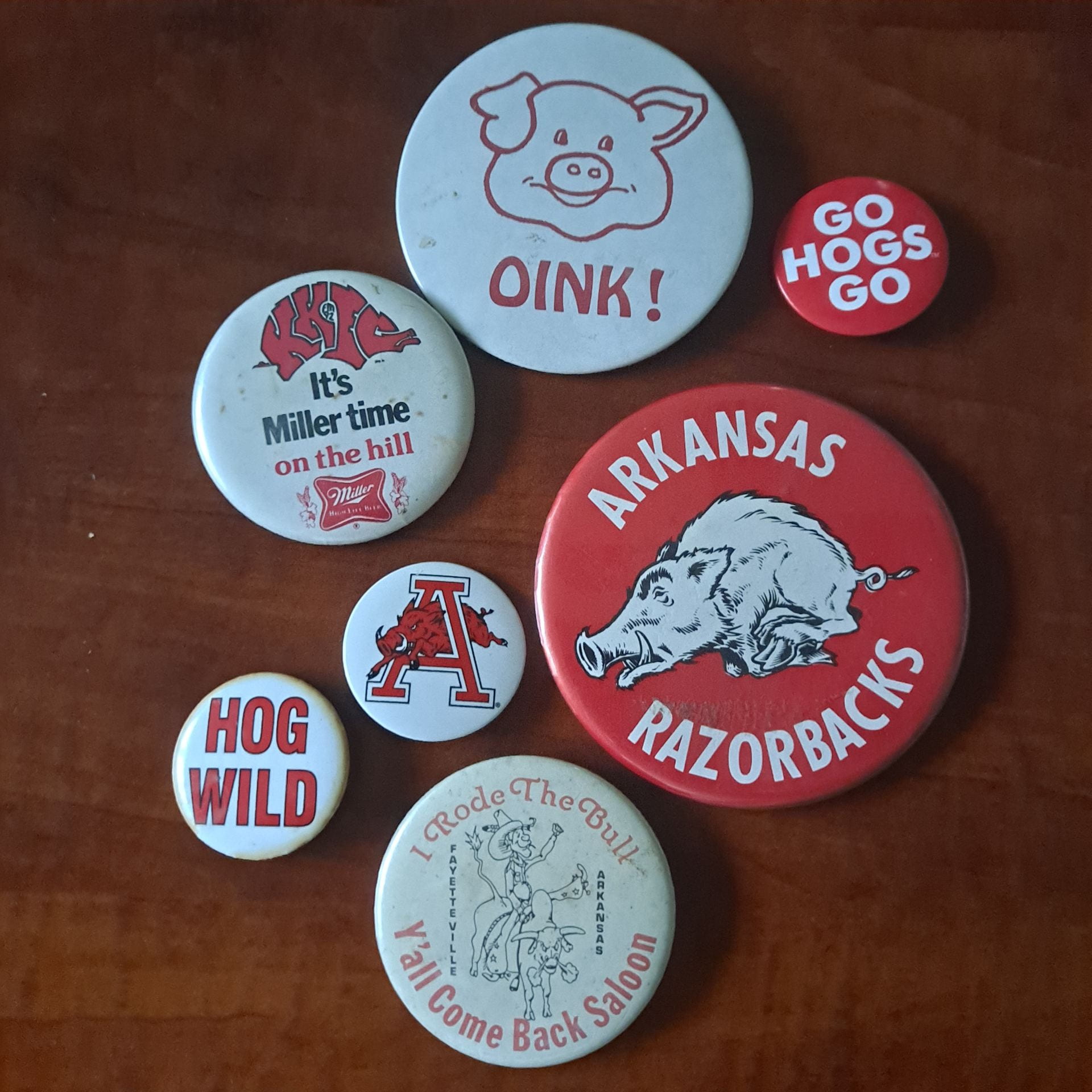 Seven Razorback-themed buttons of various sizes. 1. Small red one that says "Go hogs go". 2. A large red one with a Razorback that says "Arkansas Razorbacks." 3. A large one with a cowboy on a bull that says "I rode the bull, ya'll come back soon, Fayetteville Arkansas" 4. A small white one that says "Hog wild." 5. A small white one with a Razorback running through an A. 6. A large white one with a Razorback that says "It's Miller time on the hill" 7. A large white one with a pig face that says "oink!"