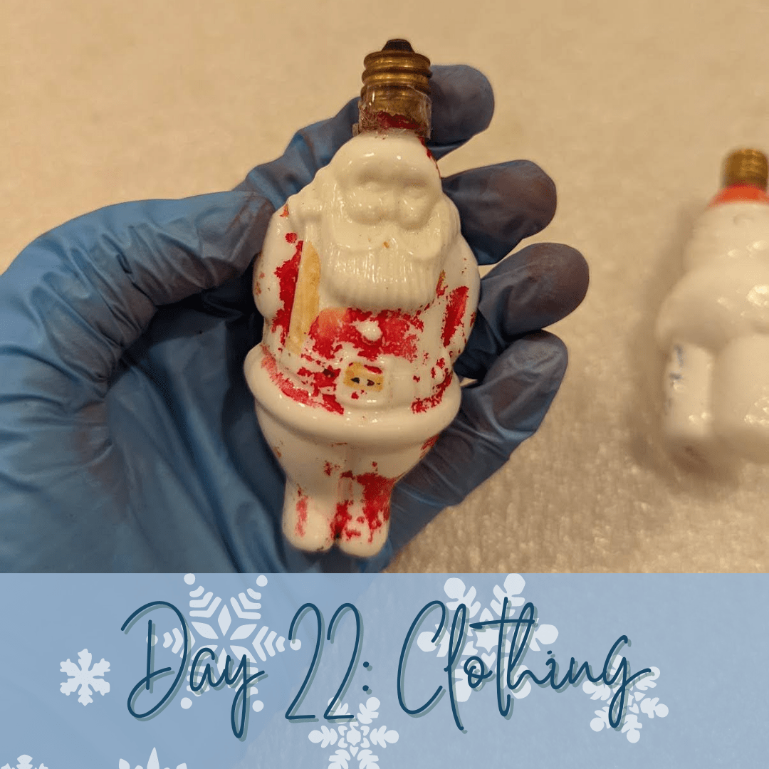 A blue nitrile gloved hand holding a Santa Claus shaped lightbulb. The red of Santa's suit is worn off quite a bit. A blue banner overlays at the bottom of the image with the words "Day 22: Clothing."