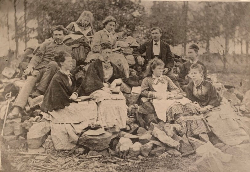 A black and white image featuring nine individuals sitting and posing on a large pile of rocks with trees in the background.