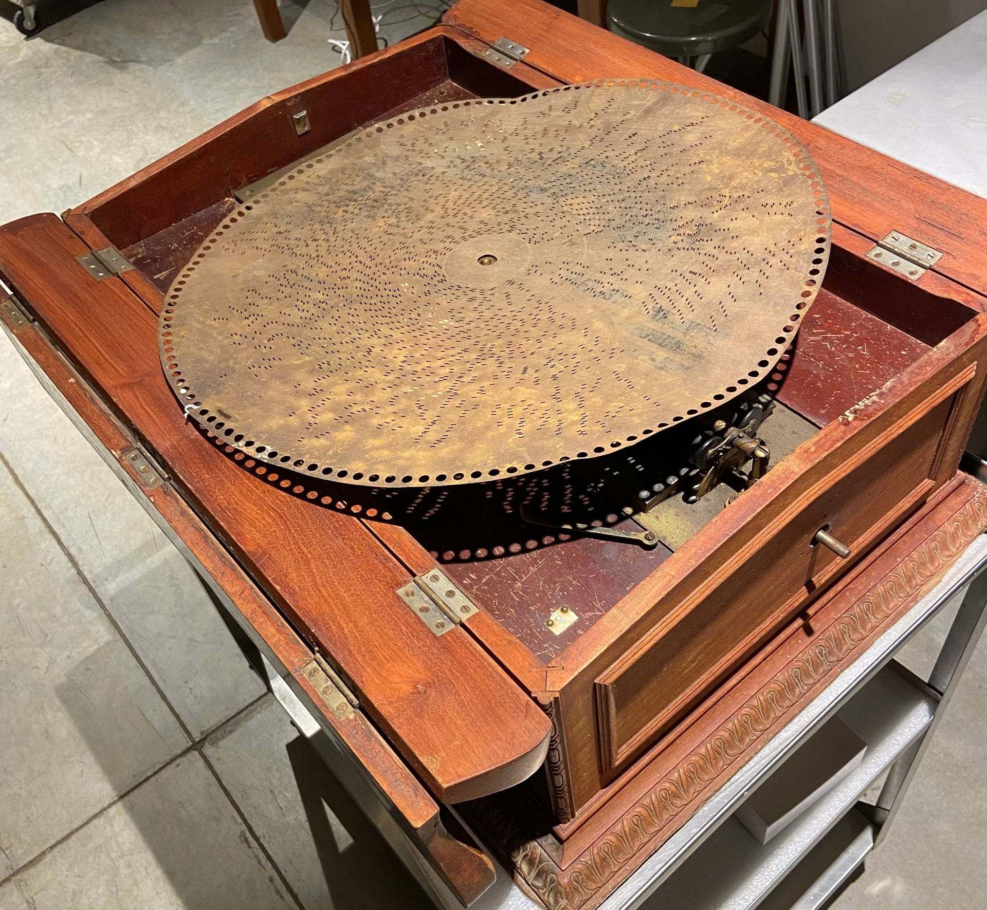 A wooden box on a metal cart. The top of the box has a lid that hinges open on two sides. It is wide open showing the interior. The interior has a large metal disk laying on top. There is a small hole in the middle where a pin from the wooden box shows through.