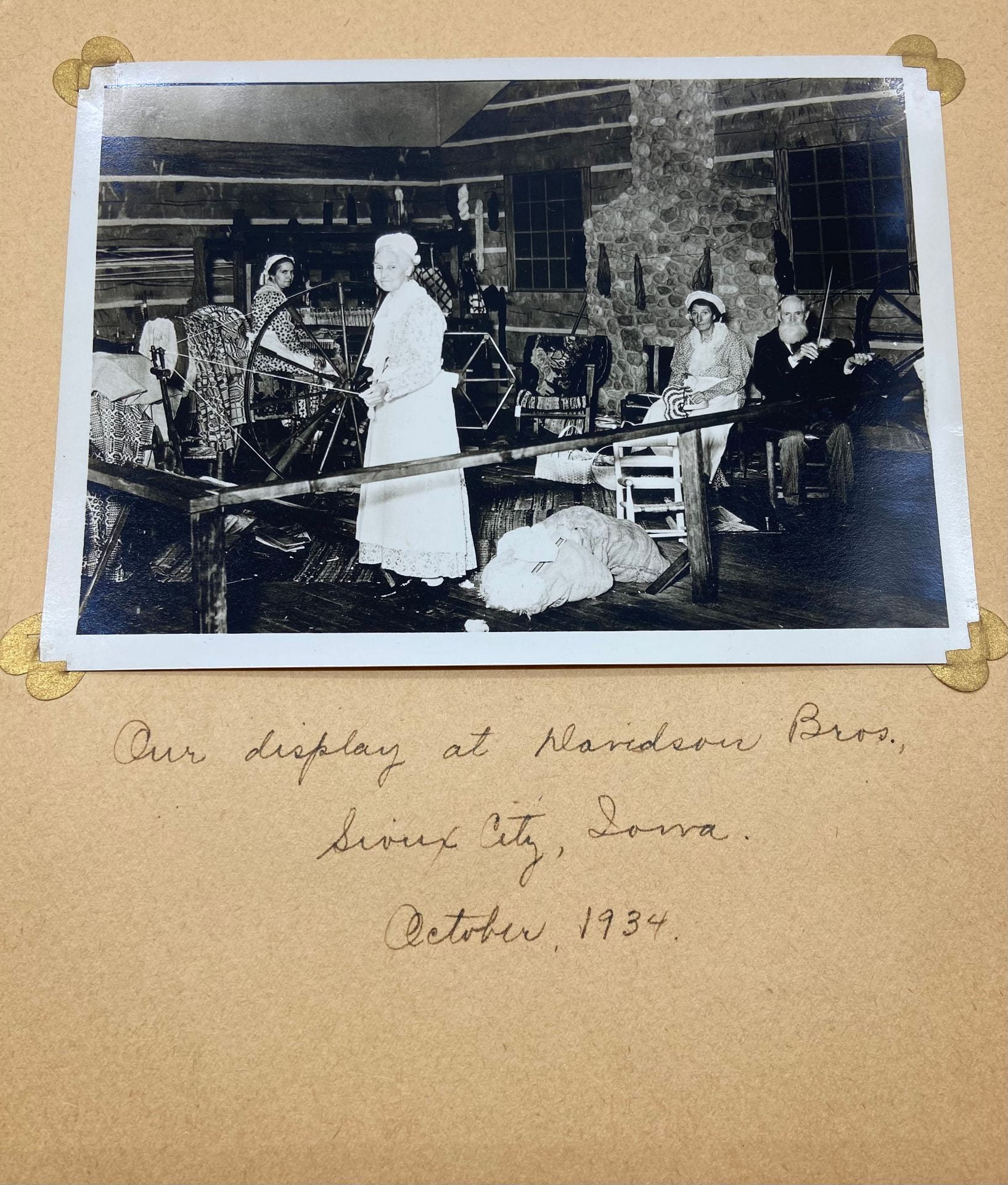A faded page with a black and white photograph adhered and an inscription that says "Our display at Davidson Bros. Sioux City, Iowa. October 1934." The black and white photo features a log cabin interior and four people posed in historical clothing around traditional spinning wheels.