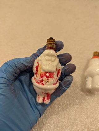 A blue nitrile gloved hand holding a Santa Claus shaped lightbulb. The red of Santa's suit is worn off quite a bit.
