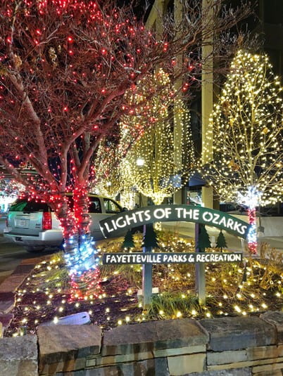 An outdoor nighttime scene featuring trees lit in red and white and a sign that says "Lights of the Ozarks Fayetteville Parks & Recreation."