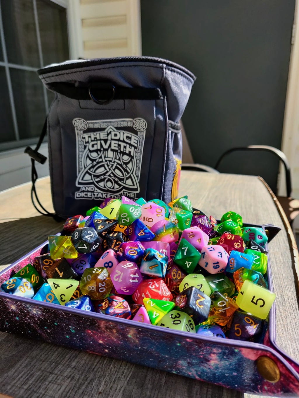 A small leather bag on a table with the words "The Dice Giveth and the Dice Taketh." A small container in front of it holds a large number of dice of all shapes and colors.