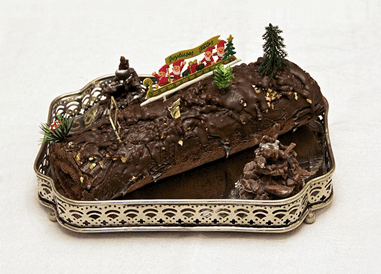 An intricately decorated dessert in the shape of a log with a four people with long white beards and red suits. The sleigh has wrapped presents in it and a flag flies above that says "Joyeuses Fetes". Small tree decorations surround it.