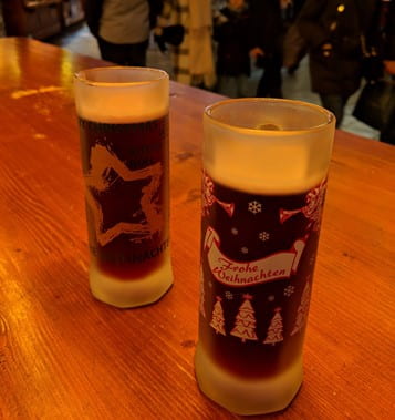Two drinks in a festive "Frohe Weihnachten" glasses set on a table with a crowd of people behind it.