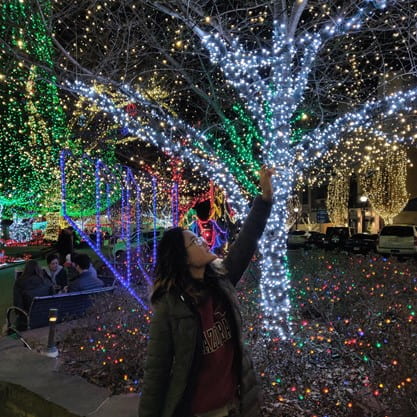 A person looking up and reaching their hand up toward a tree covered in blue lights. She's wearing a coat and it is nighttime.