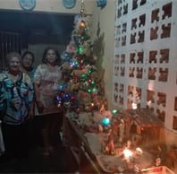 Three people poses next to each with a decorated tree to their right and a manger scene set out on a table before them.