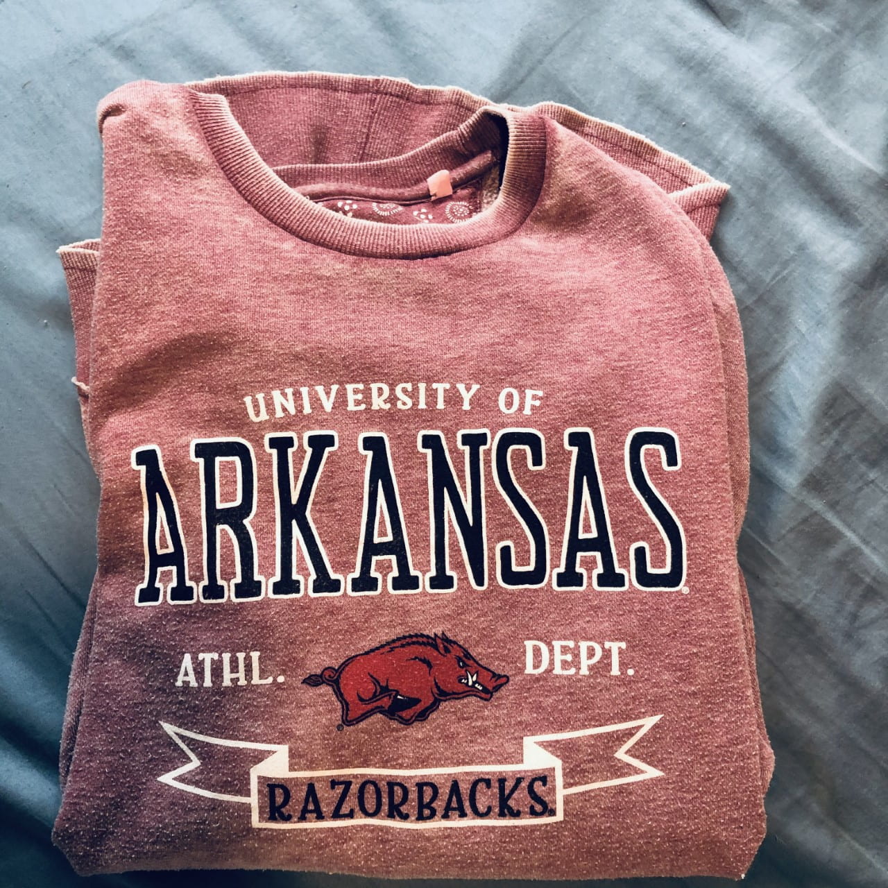 A folded light red shirt with its front facing the camera. The front has "University of Arkansas Athl. Dept. Razorbacks" with a running Razorback on it.