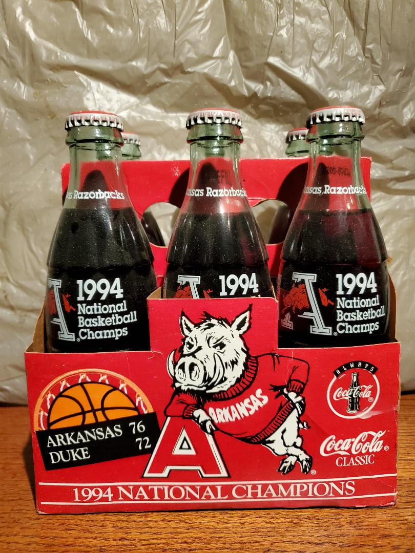 A six-pack of clear glass Coca-Cola bottles with "1994 National Basketball Champs". The bottles are in red paper bottle holder with a Razorback and "1994 National Champions) on the side..