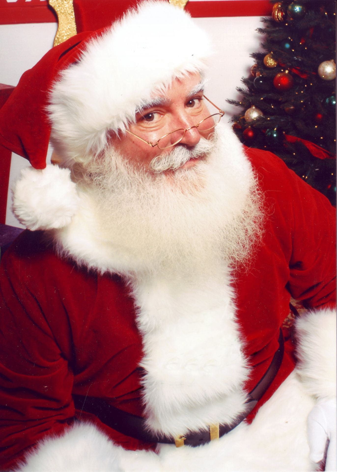 An older man with a white beard in a red suit lined with white fur. He has glasses and rosy cheeks.