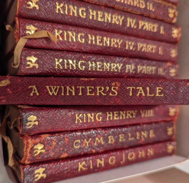 Eight red, worn book spines with gold lettering stacked horizontally. Shakespeare's "King Henry IV" is included, as well as "A Winter's Tale." The latter is pulled out and focused over the rest.