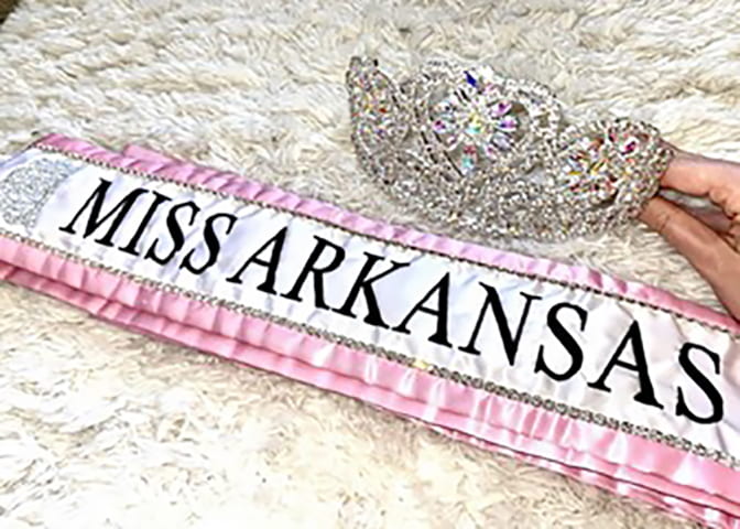 A cloth banner lined in pink with the words "Miss Arkansas" is laid out and a hand holds out an intricate crown just above it.