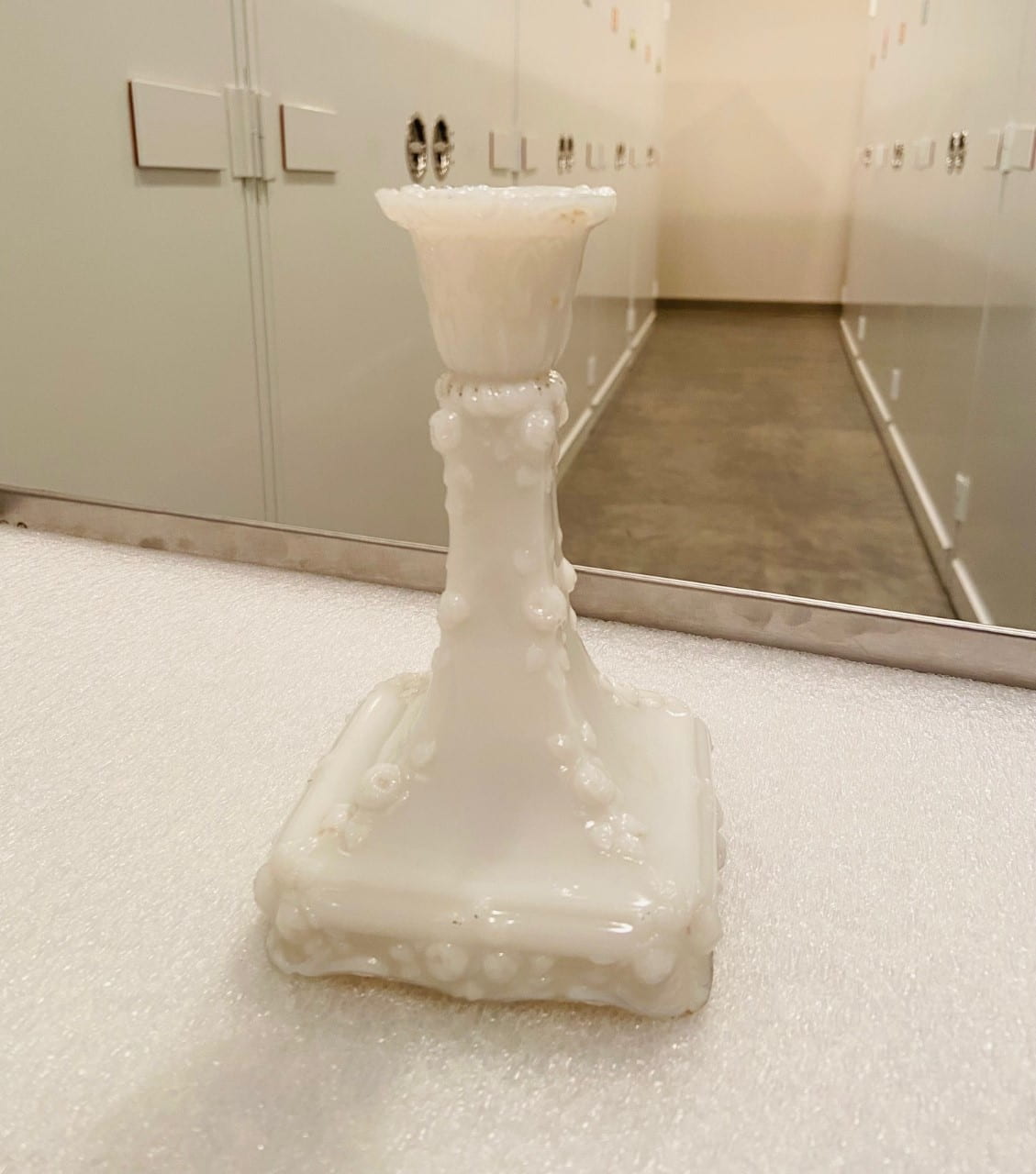 A decorative white glass candlestick on a white surface with cabinets in the background.