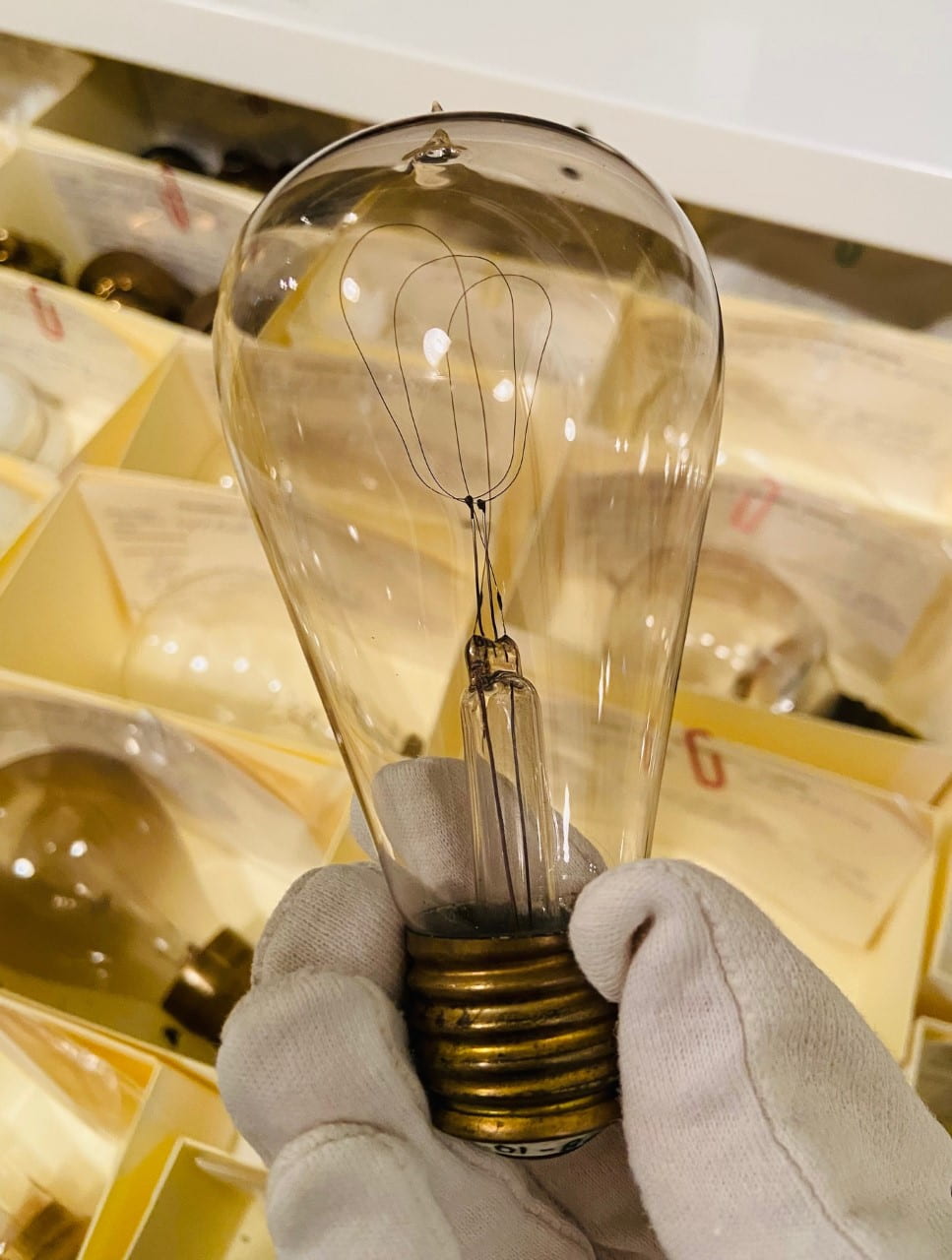 A clear lightbulb held between cotton-gloved fingers. There are more lightbulbs in small boxes behind the featured one.