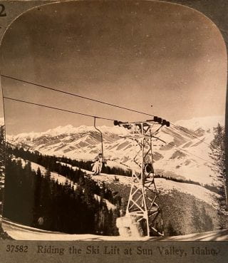 A winter scene with a person on a ski lift and mountains in the background.