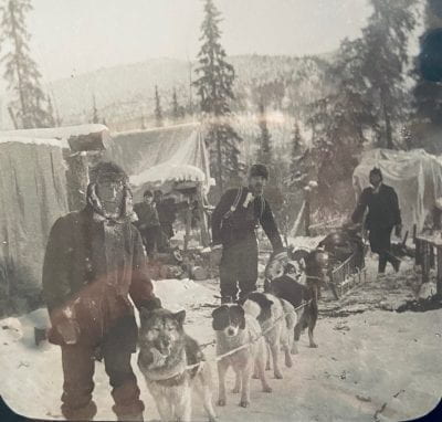 A black and white photograph of three men standing next to a dog sled team outside in the snow.