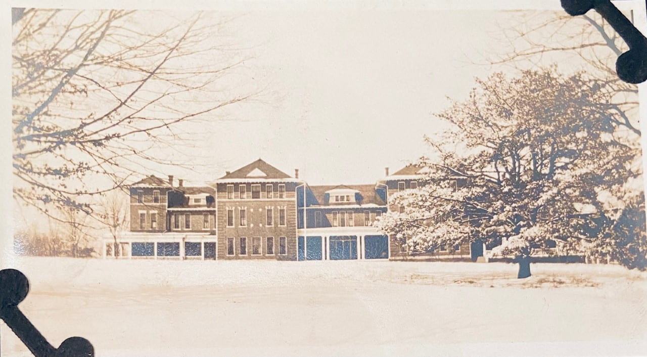A black and white photograph of a large building in the background. In the foreground is wide open outdoor area with a couple trees off to the sides. Snow blankets the ground.